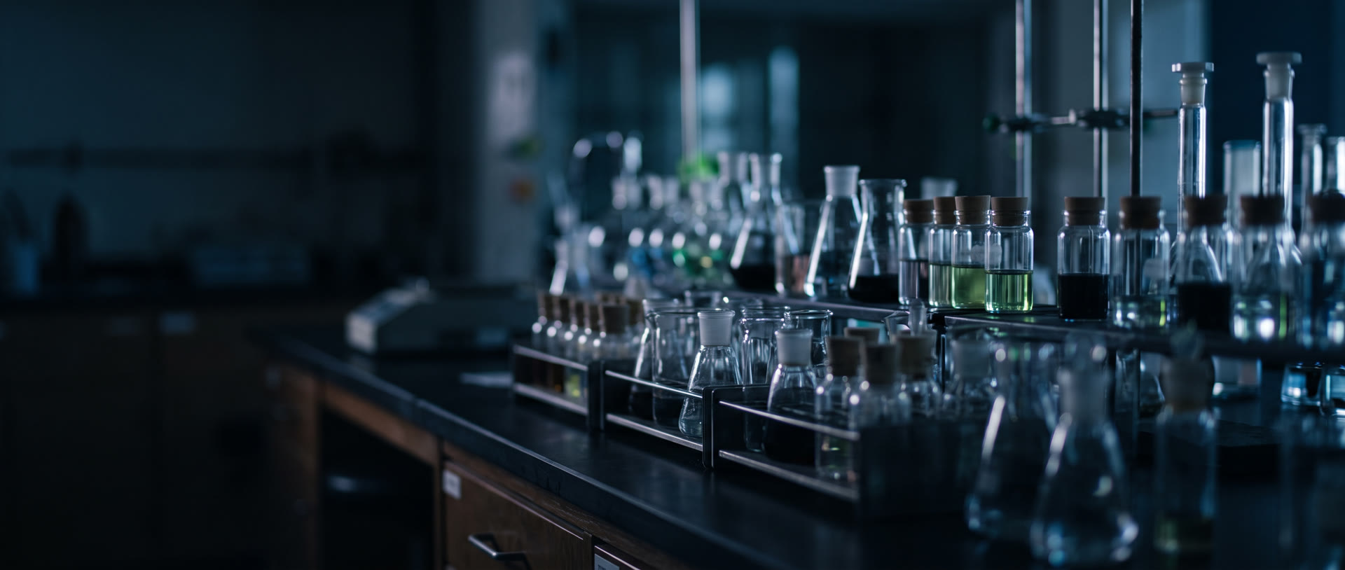 Paillasse de laboratoire dans la pénombre bleutée, rangées de flacons, d’erlenmeyers et de fioles de verre, certains remplis d’un liquide vert (décor)